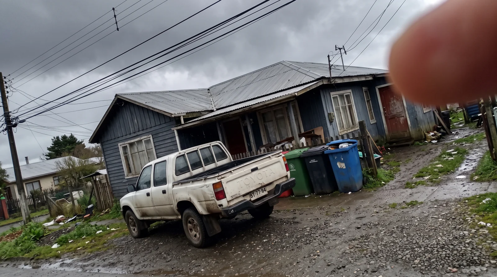 The same house shot on a grey day, with a pickup truck blocking the facade, a finger over the lens, a tilted horizon and rubbish bins in view